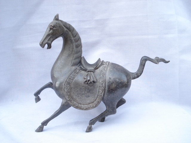 A dark metal statue of a horse in a galloping pose, featuring a saddle with decorative trim, set against a plain white…