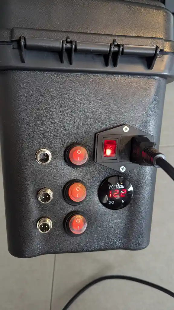 A close-up of a black, textured power panel with three red illuminated toggle switches, three metal ports, a digital voltage…