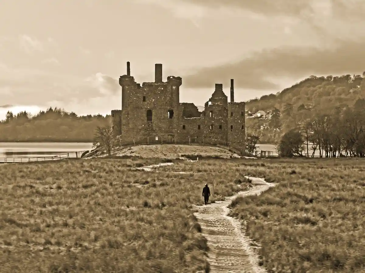 A sepia-toned photo shows a lone person walking along a muddy path toward the ruins of a large stone castle situated beside…