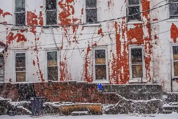 A weathered, snow-covered building with peeling red and white paint and boarded windows, with a small blue object sitting on…