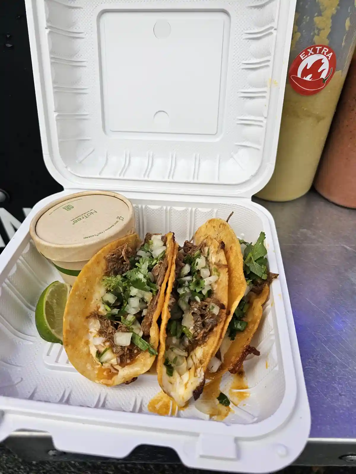 A white takeout container holds two tacos with shredded meat, chopped onions, and cilantro, next to a lime wedge and a small…