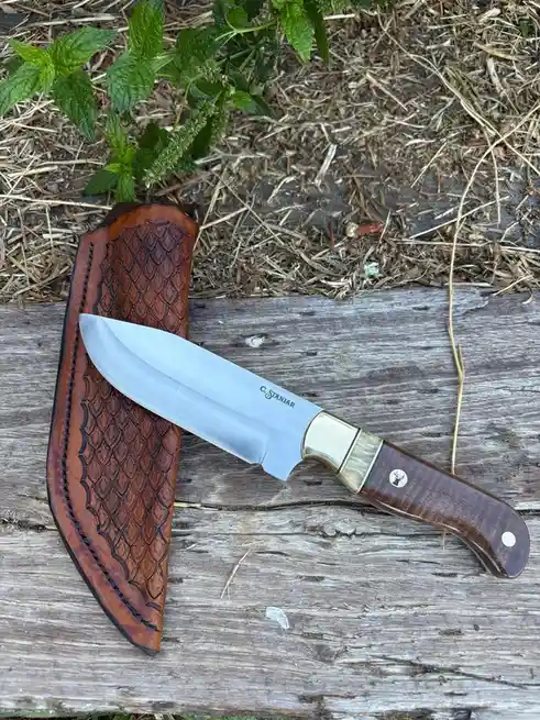 A custom-made knife with a wooden handle and brass bolster lies on weathered wood next to a brown leather sheath with a…