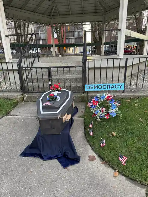 A black coffin draped in a navy blue cloth sits on a sidewalk next to a grassy area, adorned with red roses, a white cross…