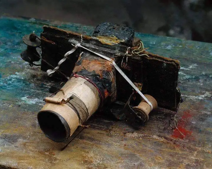 A heavily damaged, burnt, and patched-up camera lies on a rusty, weathered surface. The camera's body is charred black, its…