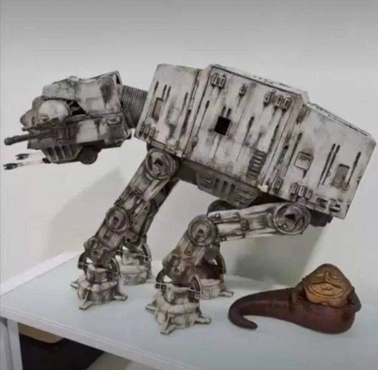 A heavily weathered, white and gray AT-AT walker model is leaning precariously on a small, brown, slug-like creature with a…