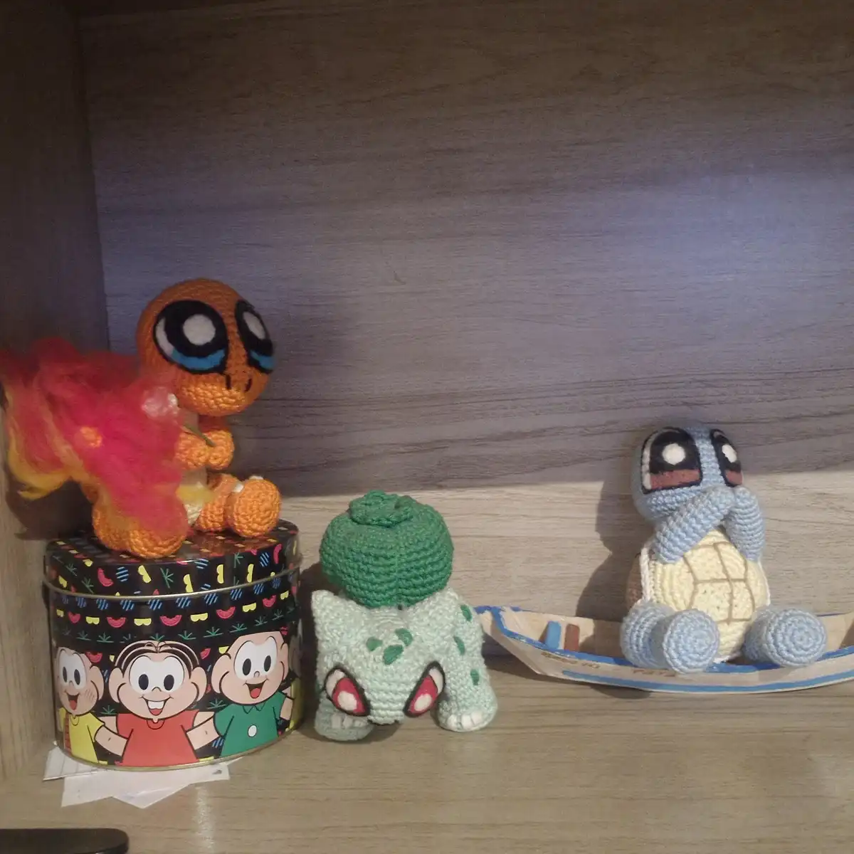 Three crocheted Pokémon—Charmander, Bulbasaur, and Squirtle—are arranged on a wooden shelf. Charmander sits atop a colorful…