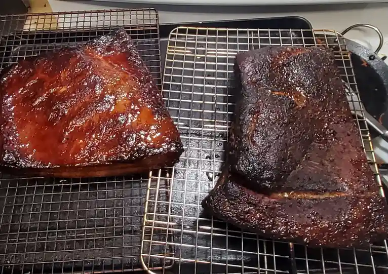 Two large pieces of smoked meat, likely ribs, sit on cooling racks. The one on the left is glossy and caramelized with a…