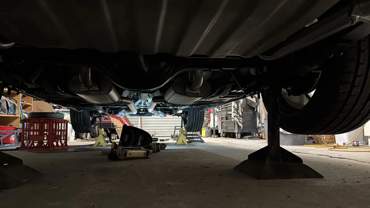 A low-angle photo looking up from under a car that's been jacked up on a lift in a garage, showing the exhaust pipes and…