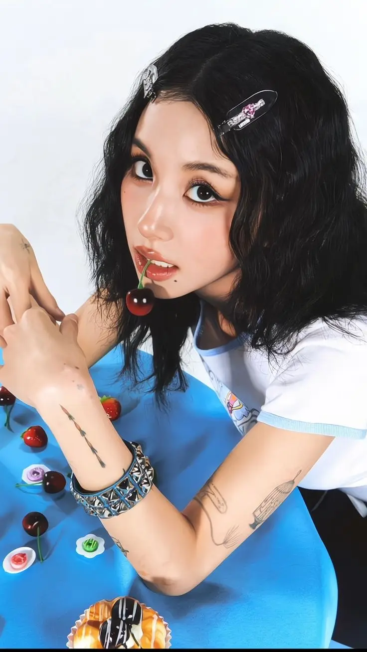 A young woman with dark wavy hair and decorative hair clips looks directly at the camera while holding a cherry by its stem…