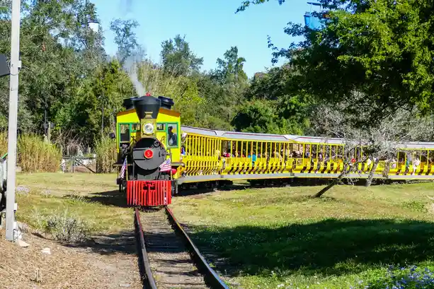 A bright yellow and red steam train with passengers in open-air cars travels along tracks through a grassy, tree-lined area…