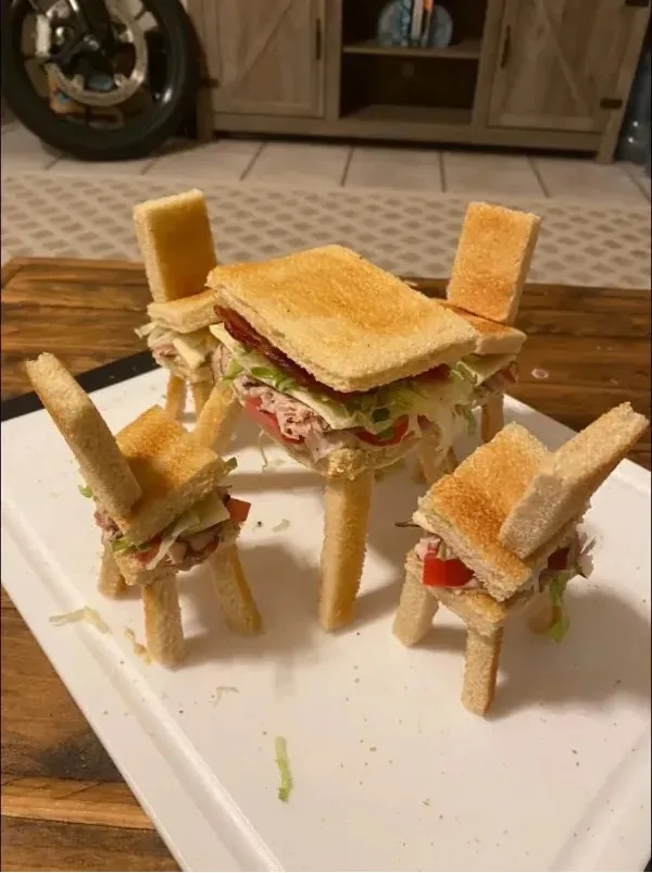 A central sandwich sits atop a tiny table made of bread, surrounded by four miniature sandwich chairs, all crafted from…