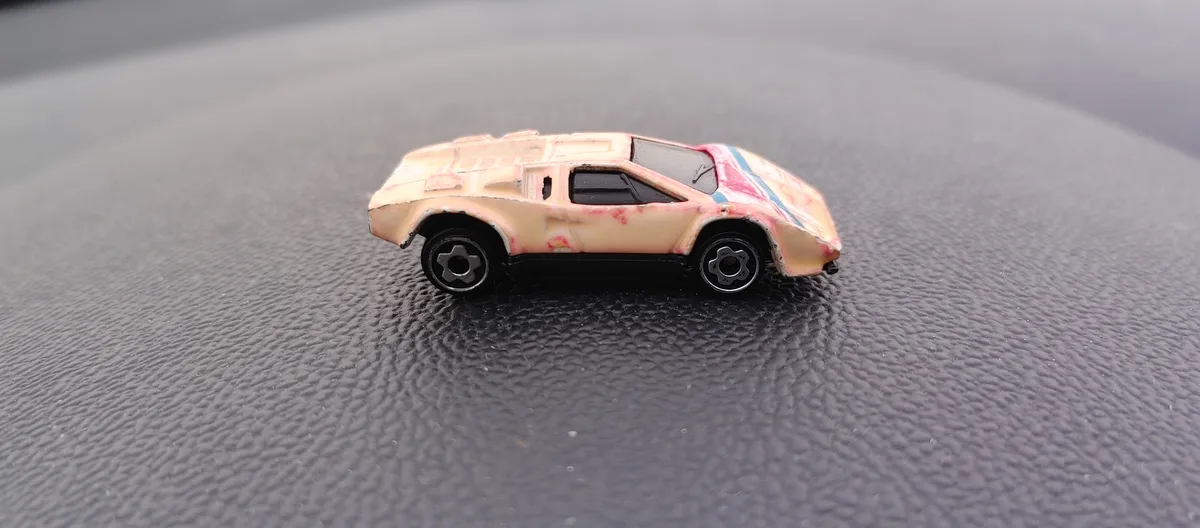 A heavily worn and chipped toy car with faded pink and blue stripes sits on a textured black surface. The overlaid text…