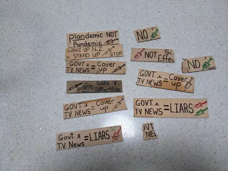 Multiple handmade cardboard signs with handwritten anti-vaccine and anti-government messages are scattered on a speckled…