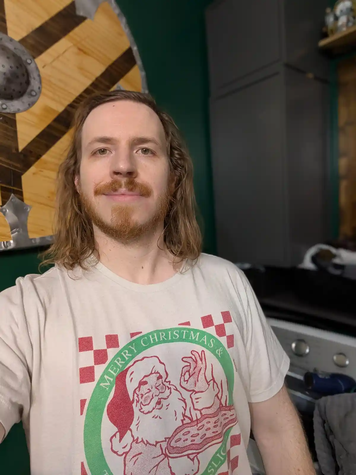 A man with long hair and a reddish beard wears a t-shirt with a graphic of Santa Claus holding a pizza slice and the text…