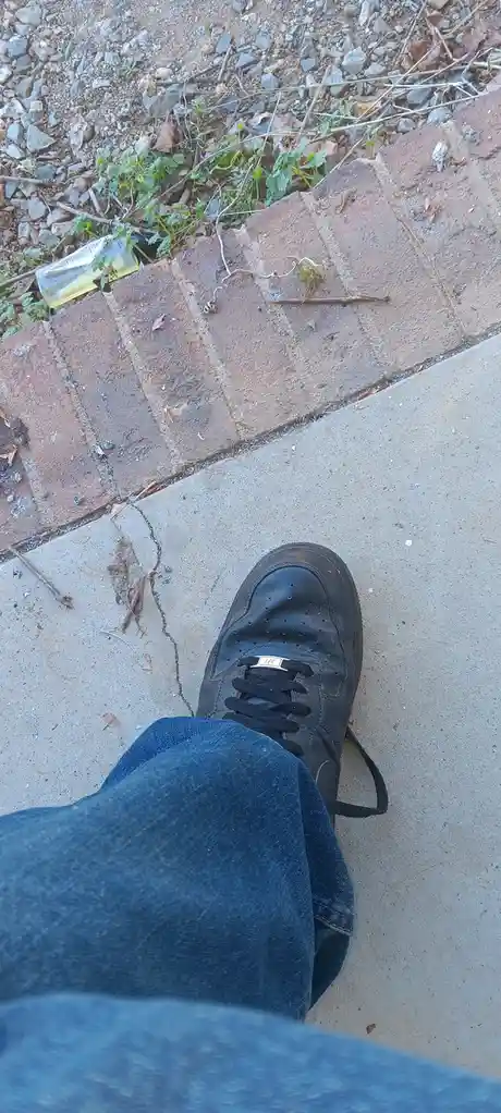 A first-person view shows a person's foot in a black leather shoe and blue jeans standing on a concrete sidewalk next to a…