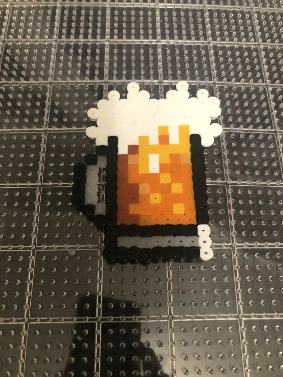 A pixel-art style beer mug made from perler beads sits on a clear pegboard, featuring a black mug with orange and yellow…