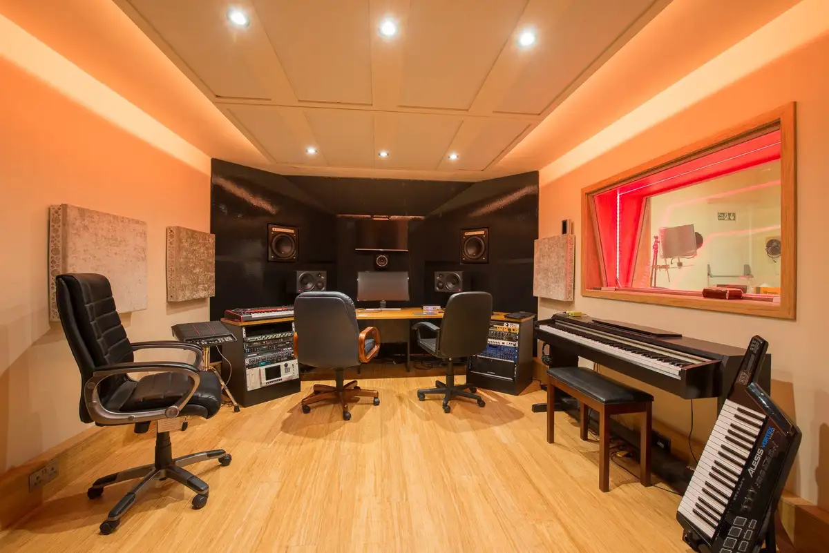 A professionally equipped music studio with soundproofing, multiple chairs, a mixing desk, speakers, a piano, and a…