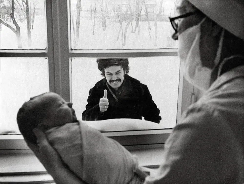 A black-and-white photo shows a man with a mustache giving a thumbs-up gesture through a window while a newborn baby is…