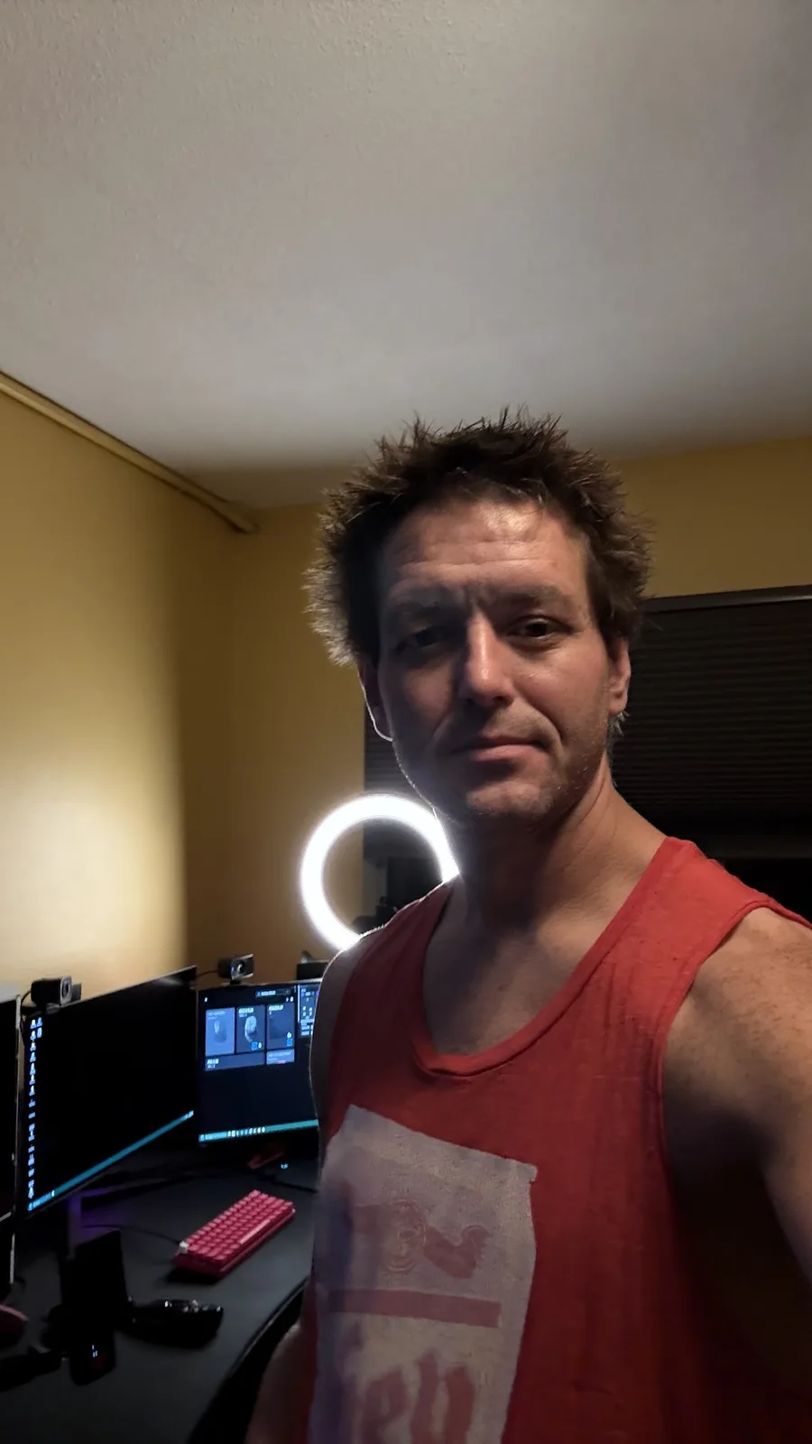 A man with messy hair and a red tank top takes a selfie in a dimly lit room with a ring light behind him and multiple…