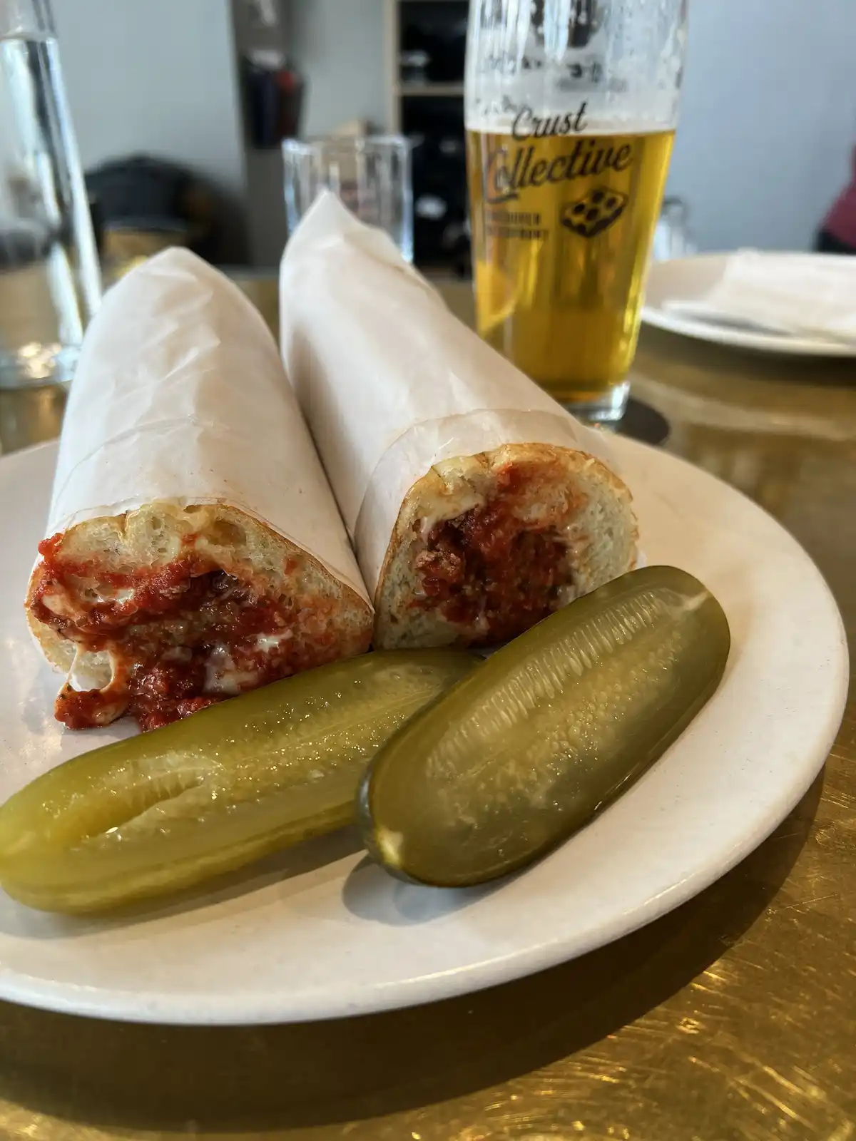 A plate holds a meatball sub cut in half, wrapped in paper, revealing meat sauce and cheese inside, alongside two pickle…