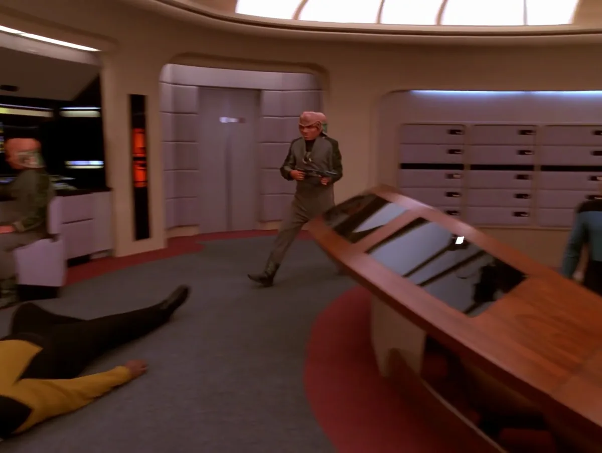 A still from Star Trek: The Next Generation shows a character in a grey uniform holding a phaser while walking past a fallen…