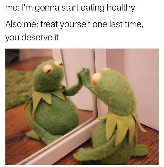 A green felt Kermit the Frog plush toy sits on a wooden floor, facing a mirror and giving a high-five to his reflection. The…
