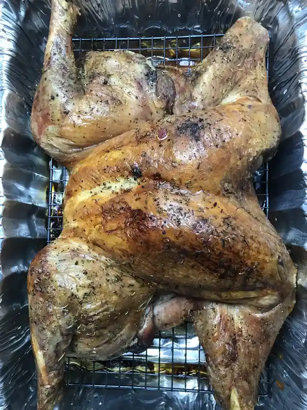 A roasted chicken sits on a wire rack inside a foil-lined baking pan, its skin golden-brown and seasoned with herbs and…