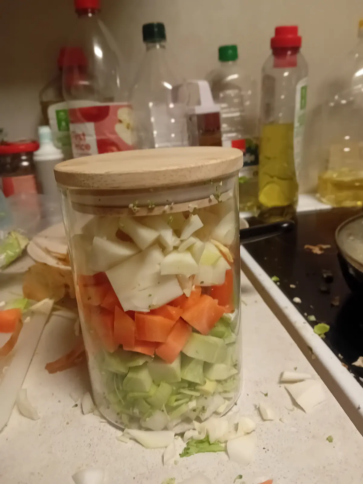 A clear glass jar with a wooden lid is filled with neatly layered, chopped vegetables including green cabbage, orange…