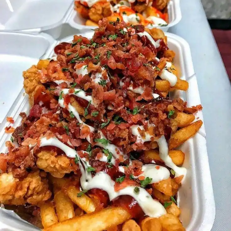 A close-up photo of a white styrofoam container filled with golden french fries topped with fried chicken pieces, crispy…