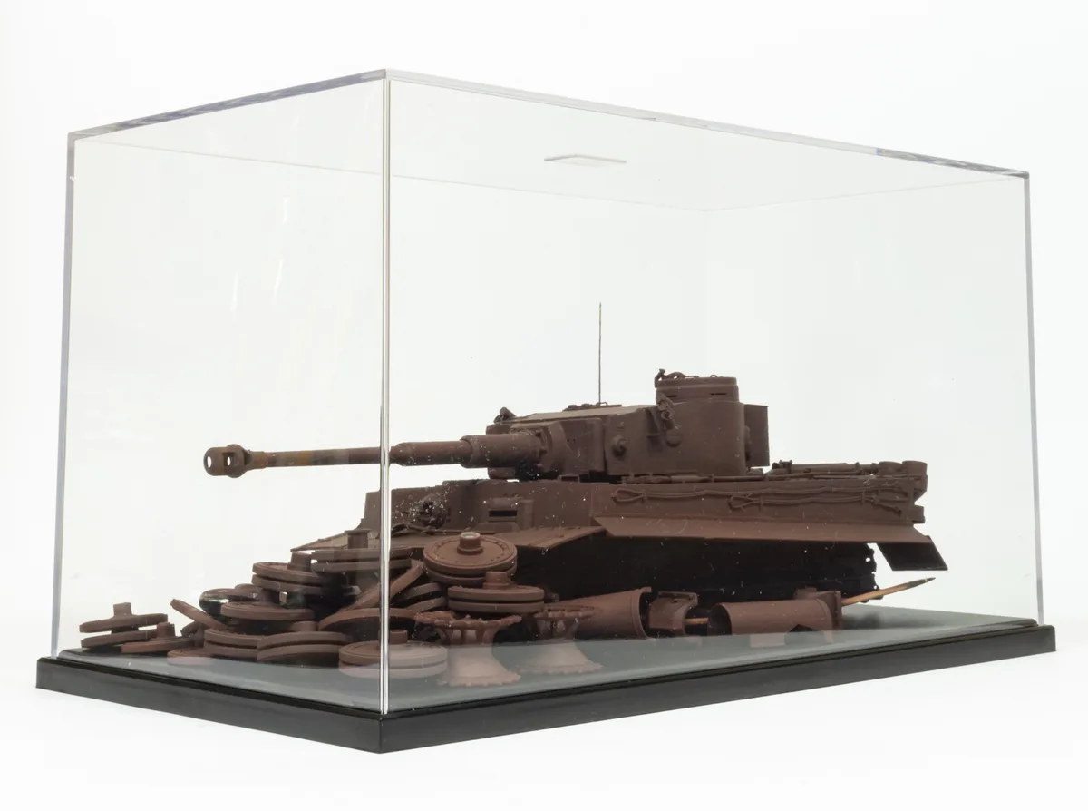 A chocolate model of a tank is displayed inside a clear acrylic case, with its tracks and wheels made of chocolate pieces…