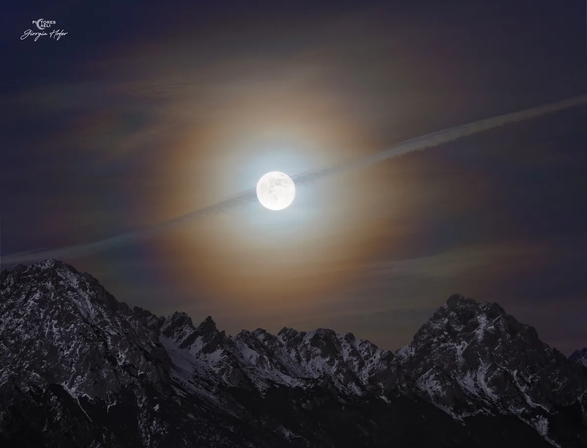 A scenic night photograph shows a full moon glowing brightly above a jagged, snow-dusted mountain range under a dark, cloudy…