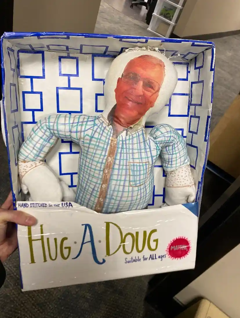 A person holds a cardboard box containing a plush doll with the face of an older man wearing glasses and a plaid shirt. The…