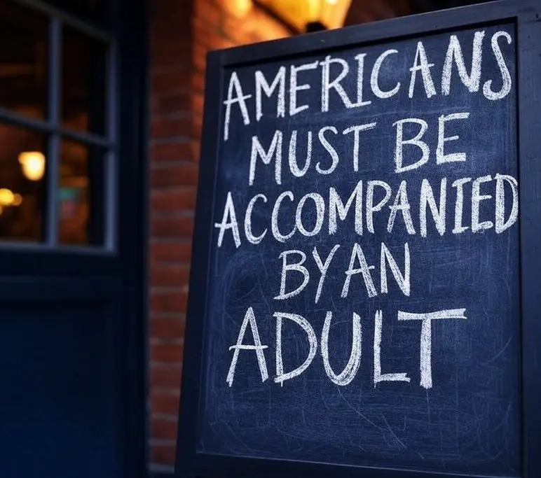 A chalkboard sign outside a brick building reads 'AMERICANS MUST BE ACCOMPANIED BY AN ADULT' in white chalk. The sign is…