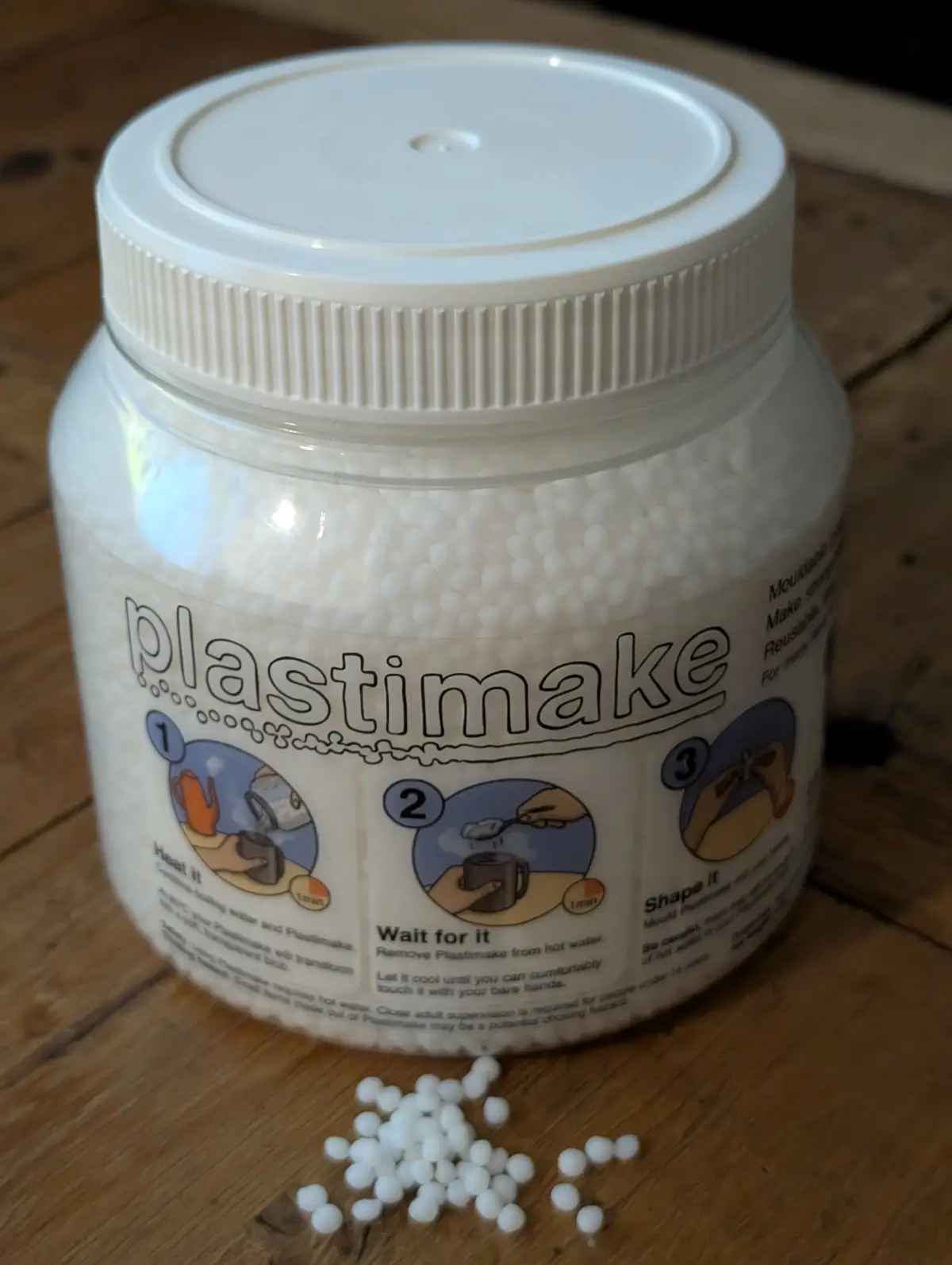 A clear plastic jar with a white lid filled with white spherical beads labeled 'plastimake' sits on a wooden surface, with a…