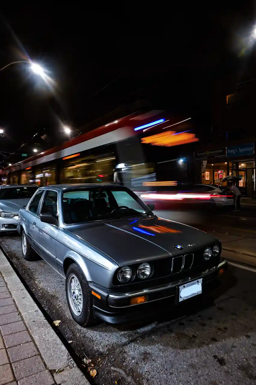 A silver 1980s BMW 3-Series sedan is parked on a wet city street at night, sharply in focus while blurred motion streaks…