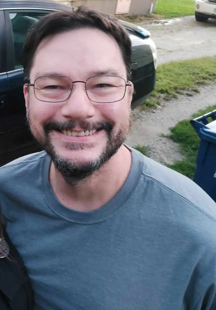 A man with glasses and a beard smiles broadly at the camera while standing outdoors near a car and a blue trash bin. The…