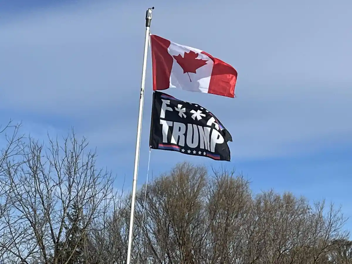 A Canadian flag flies above a black flag with white text reading 'F**K TRUMP' on a flagpole against a blue sky with bare…