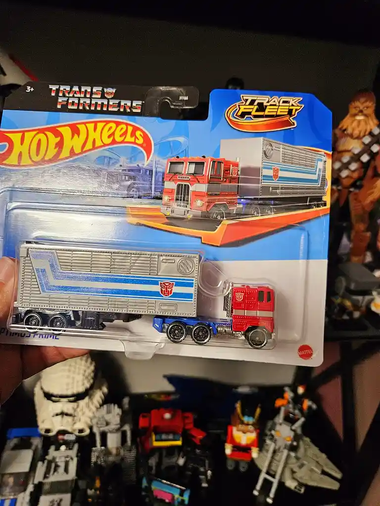 A hand holds a Hot Wheels Transformers Track Fleet toy truck, specifically Optimus Prime, in its original blister packaging…
