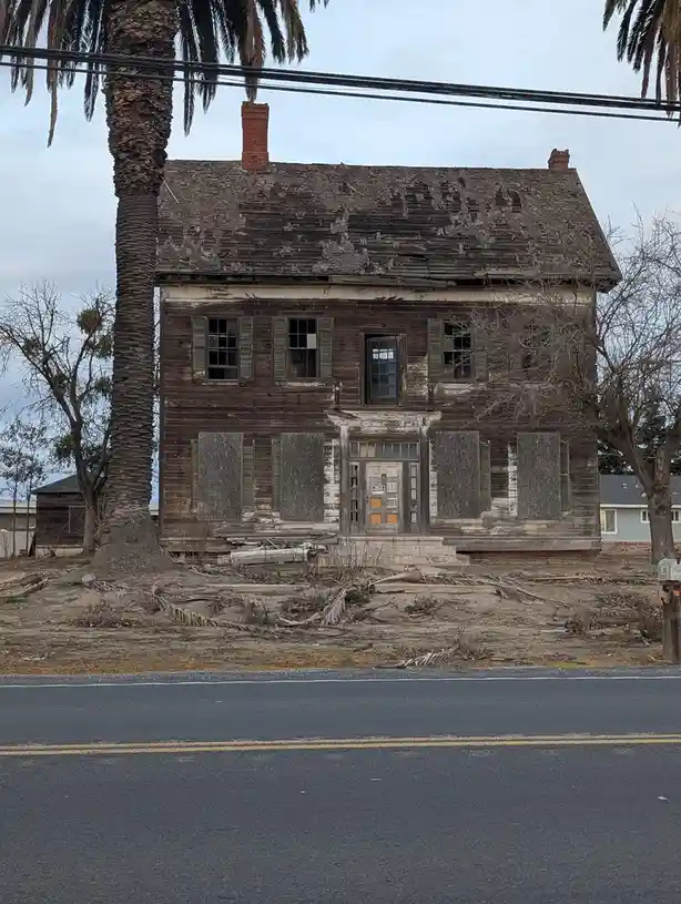 A two-story wooden house with a severely damaged roof, boarded-up windows, and peeling paint stands abandoned beside a road…