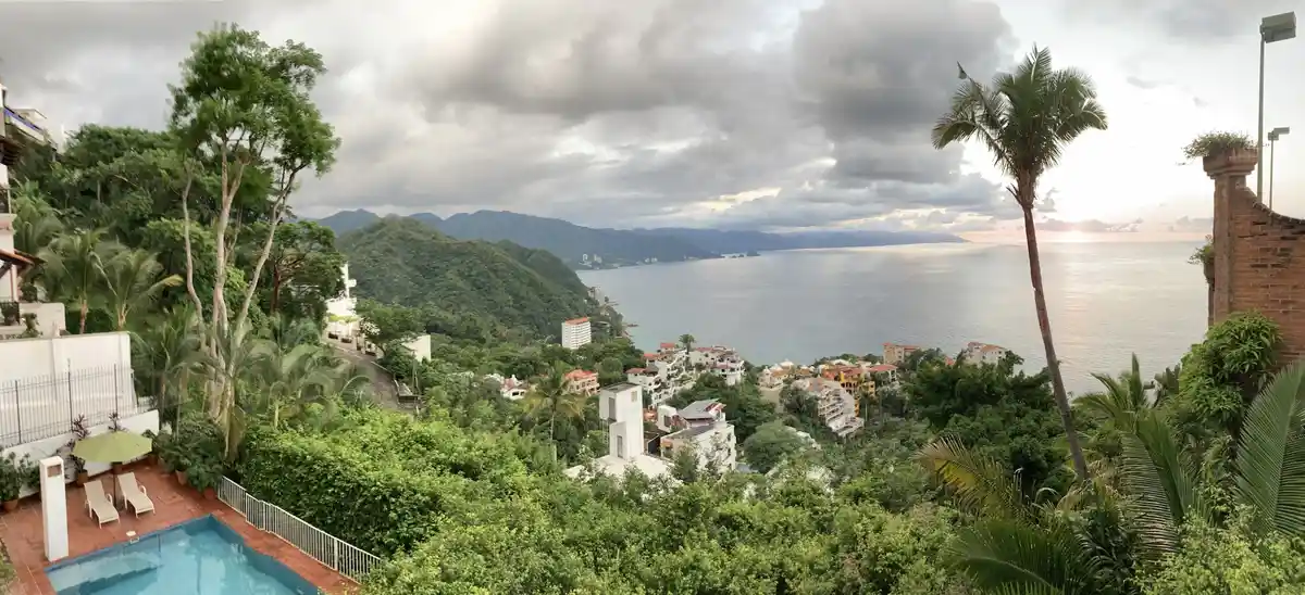 A scenic panoramic photo shows a luxurious tropical villa with a pool overlooking a lush green hillside and a vast ocean…