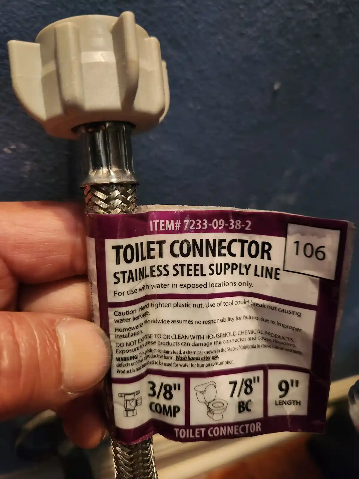 A hand holds a new toilet supply line connector with its label visible, showing specifications like 3/8" COMP, 7/8" BC, and…