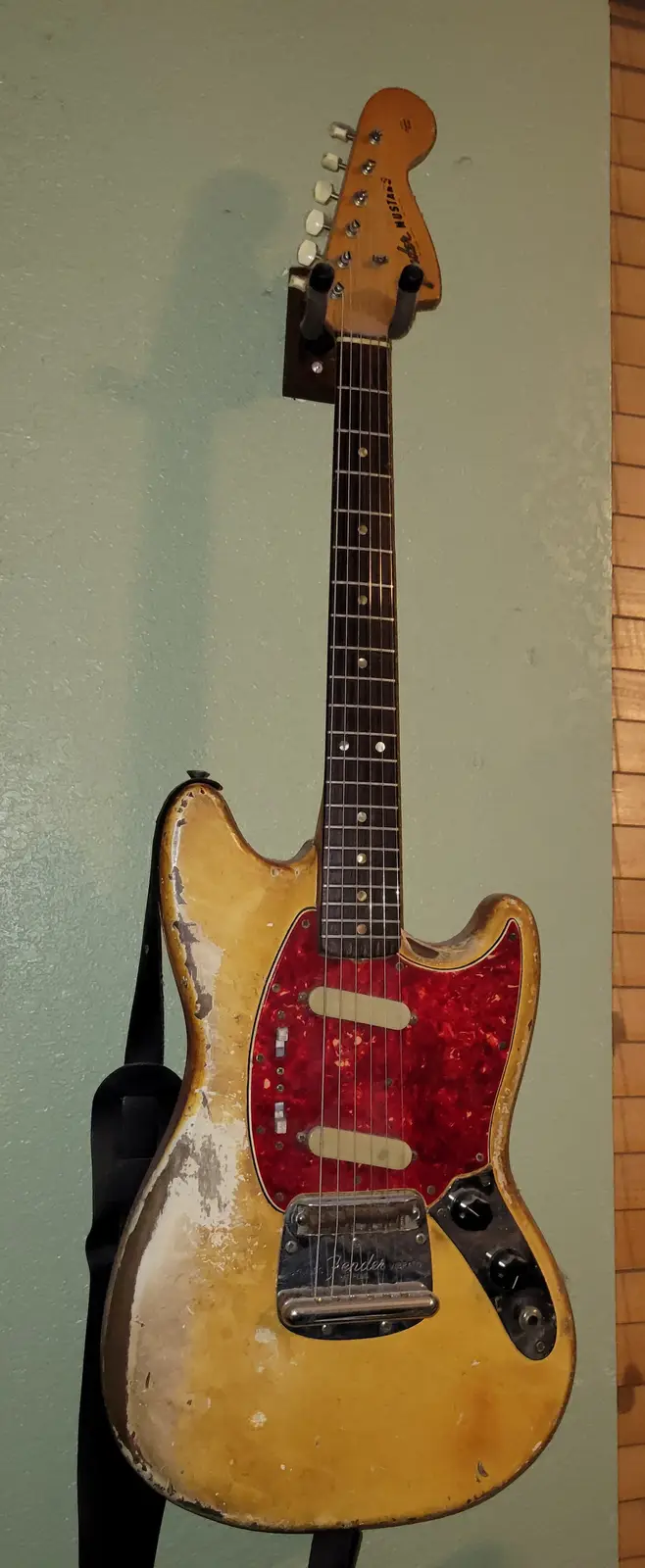 A heavily worn, vintage Fender Mustang electric guitar with a faded blonde finish and red pickguard hangs on a green wall…
