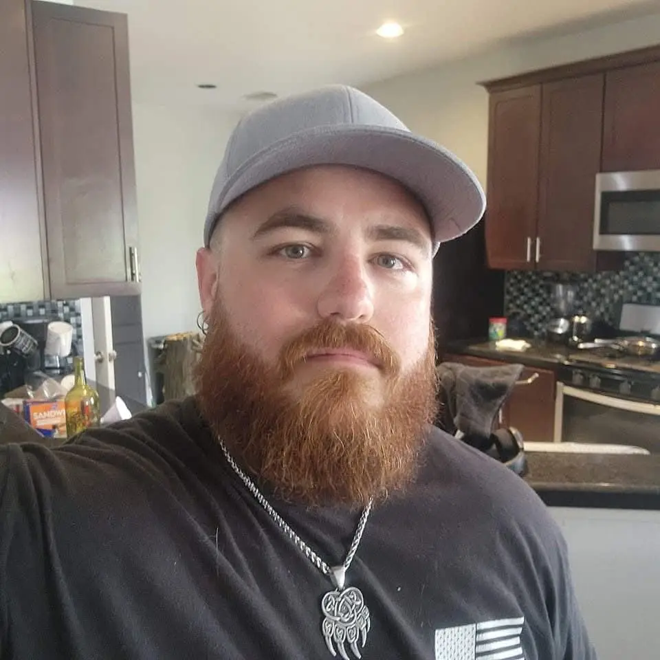 A man with a full red beard and a grey baseball cap takes a selfie in a kitchen. He wears a black t-shirt with a small…