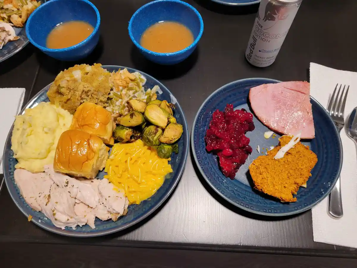 A table set with two plates of Thanksgiving food including turkey, stuffing, mashed potatoes, rolls, macaroni, Brussels…
