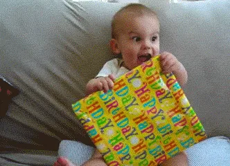 A baby sits on a couch holding a colorful 'Happy Birthday' gift wrapped present, showing escalating expressions of pure joy…