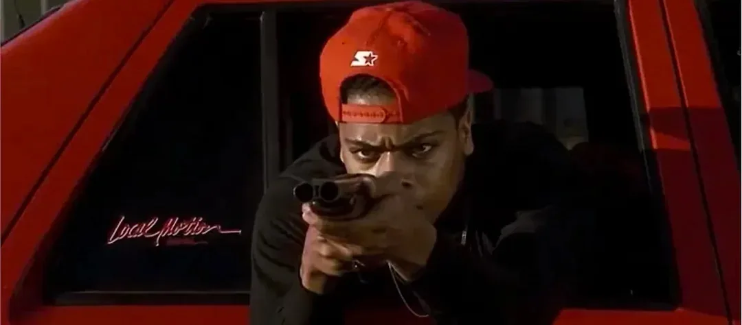 A man in a red baseball cap peers out of a red car window, holding a shotgun pointed forward with a serious, intense…