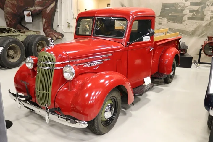 A shiny red vintage truck, likely a 1930s model, is displayed in a museum with a large dinosaur model visible in the…