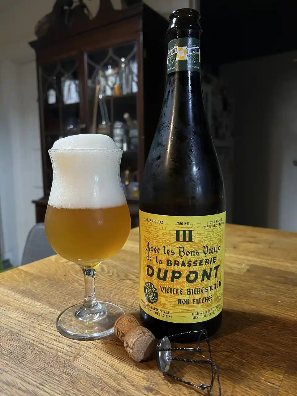 A bottle of Dupont III Belgian Saison Ale sits next to a tulip glass filled with the beer and topped with a thick white…