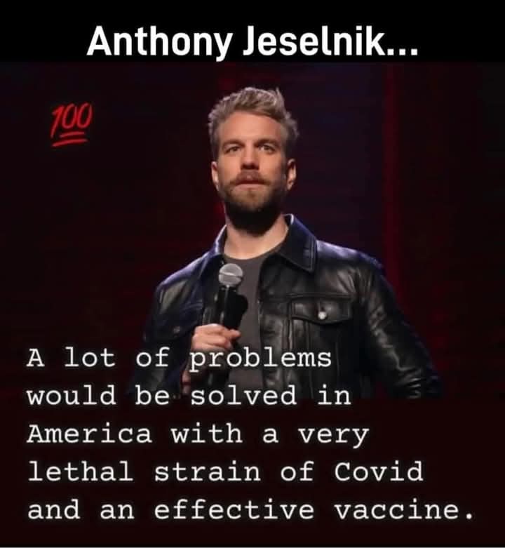 A man with short blond hair and a beard, wearing a black leather jacket over a gray shirt, stands on a stage holding a…