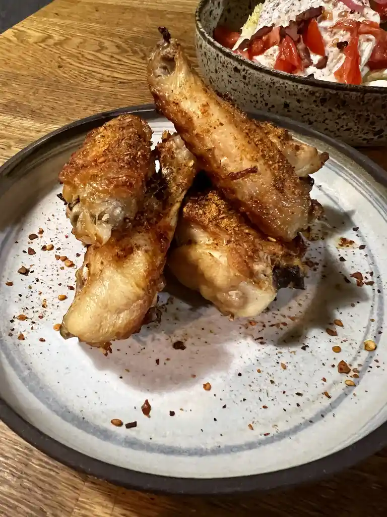 A close-up photo shows a plate with three fried chicken wings, seasoned with spices, sitting on a speckled ceramic plate. In…